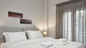 Un lit bien fait, avec du linge de lit et des serviettes blanches, une tête de lit, deux œuvres d’art accrochées au mur, une lampe de chevet et une fenêtre avec des rideaux.