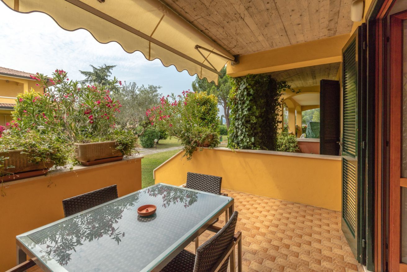An outdoor patio with a glass table and chairs, surrounded by potted plants and a yellow wall.