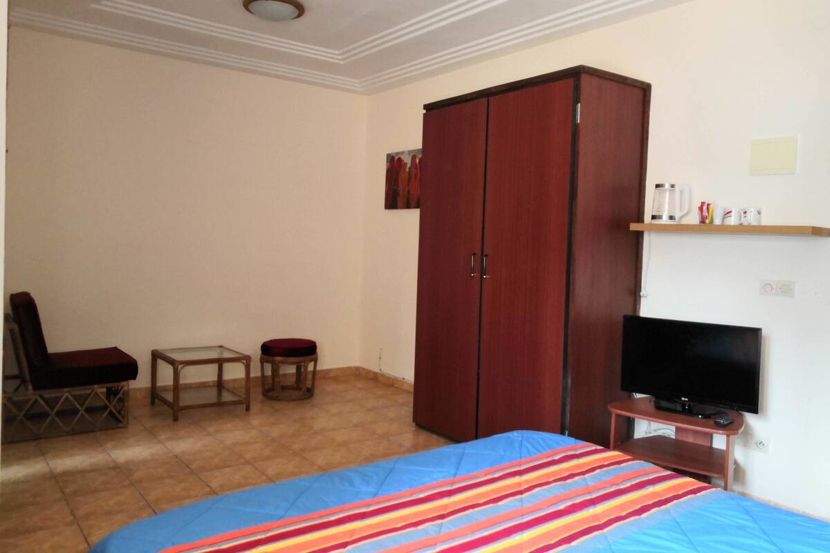 A bedroom with a bed, a TV, a wardrobe, a chair, a small table, and a shelf with items.