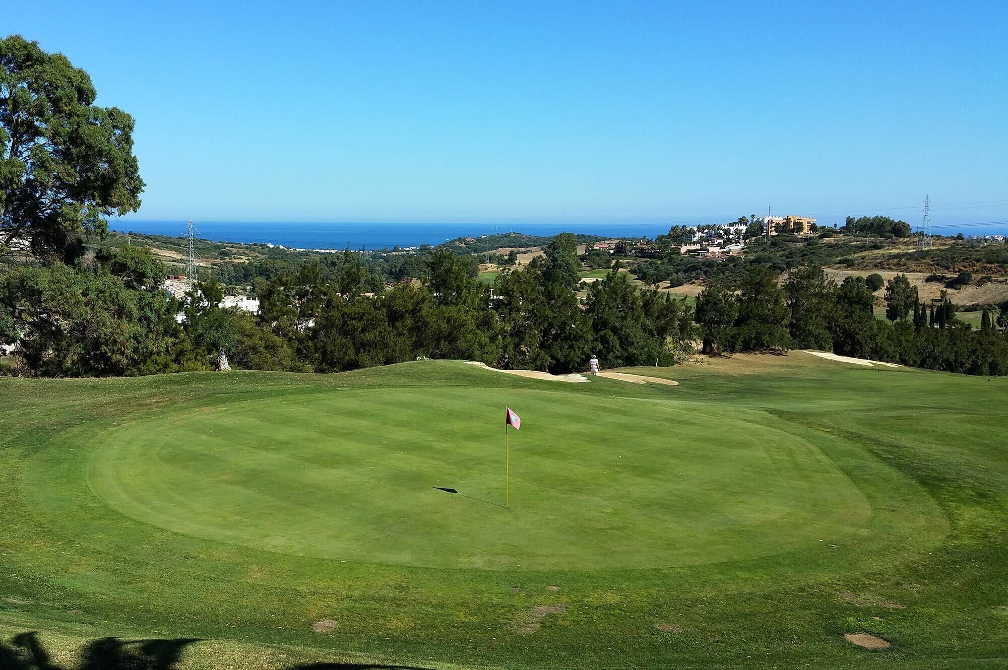 Ein Golfplatz mit Fahnenstange in der Mitte, umgeben von üppiger Vegetation und freiem Blick auf den Ozean im Hintergrund.