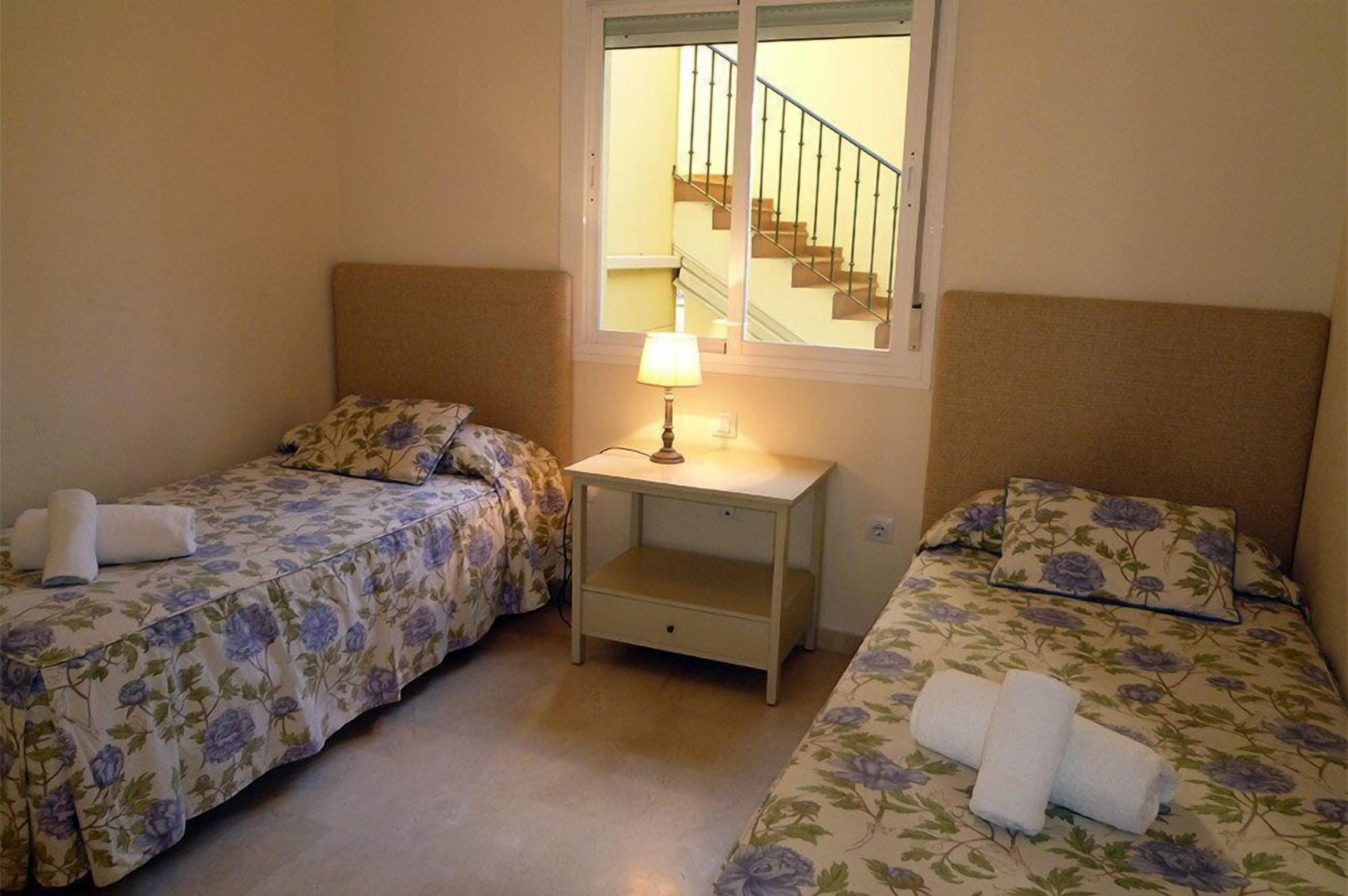 Une petite chambre propre, avec deux lits simples, une table de chevet avec une lampe et une fenêtre donnant sur un escalier.
