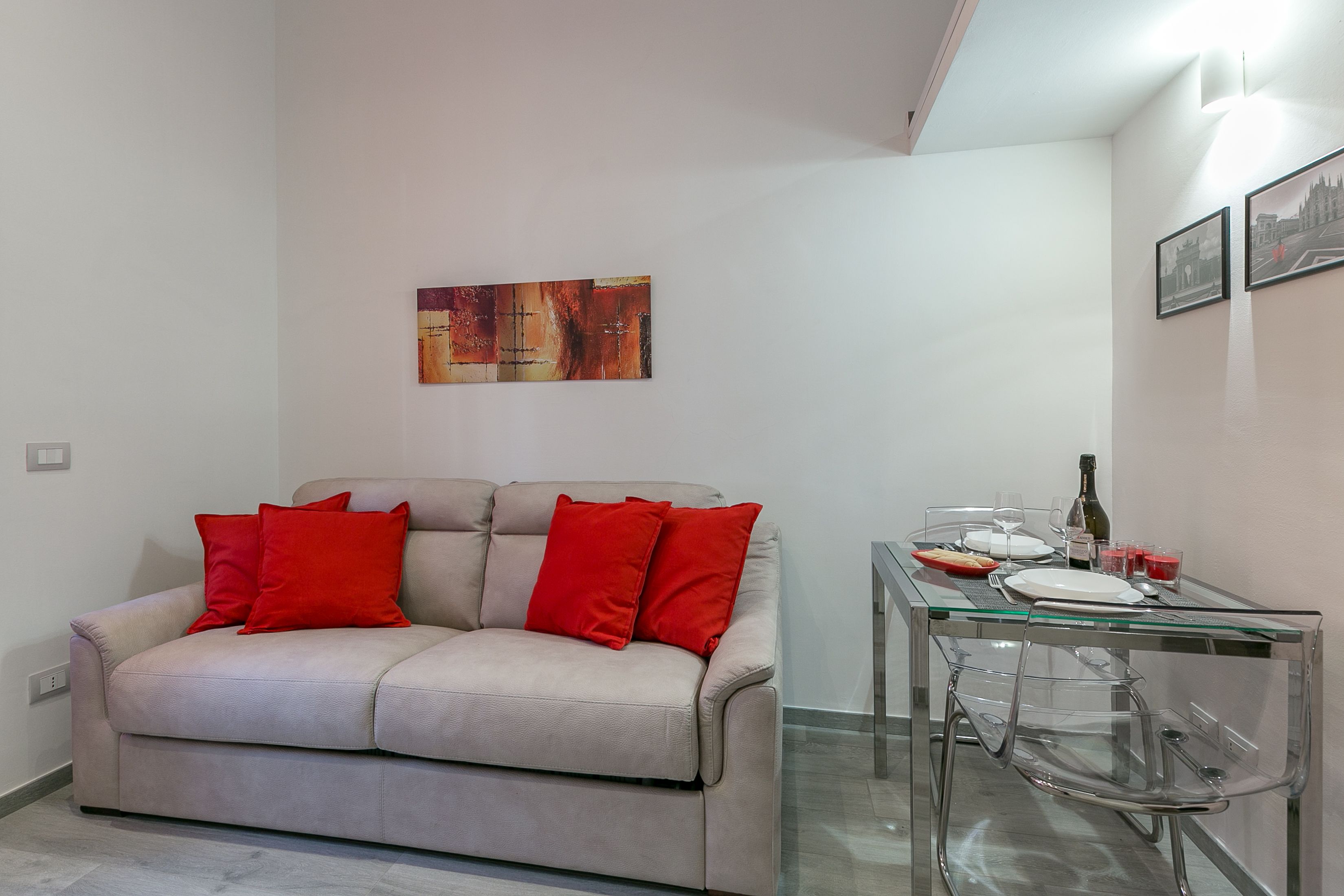 Un salon avec un canapé beige, des coussins rouges, une table à manger en verre et un tableau accroché au mur.