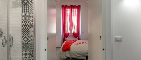 Een slaapkamer met een douche, behangtegels met een patroon, een bed met een rode deken en rode gordijnen.
