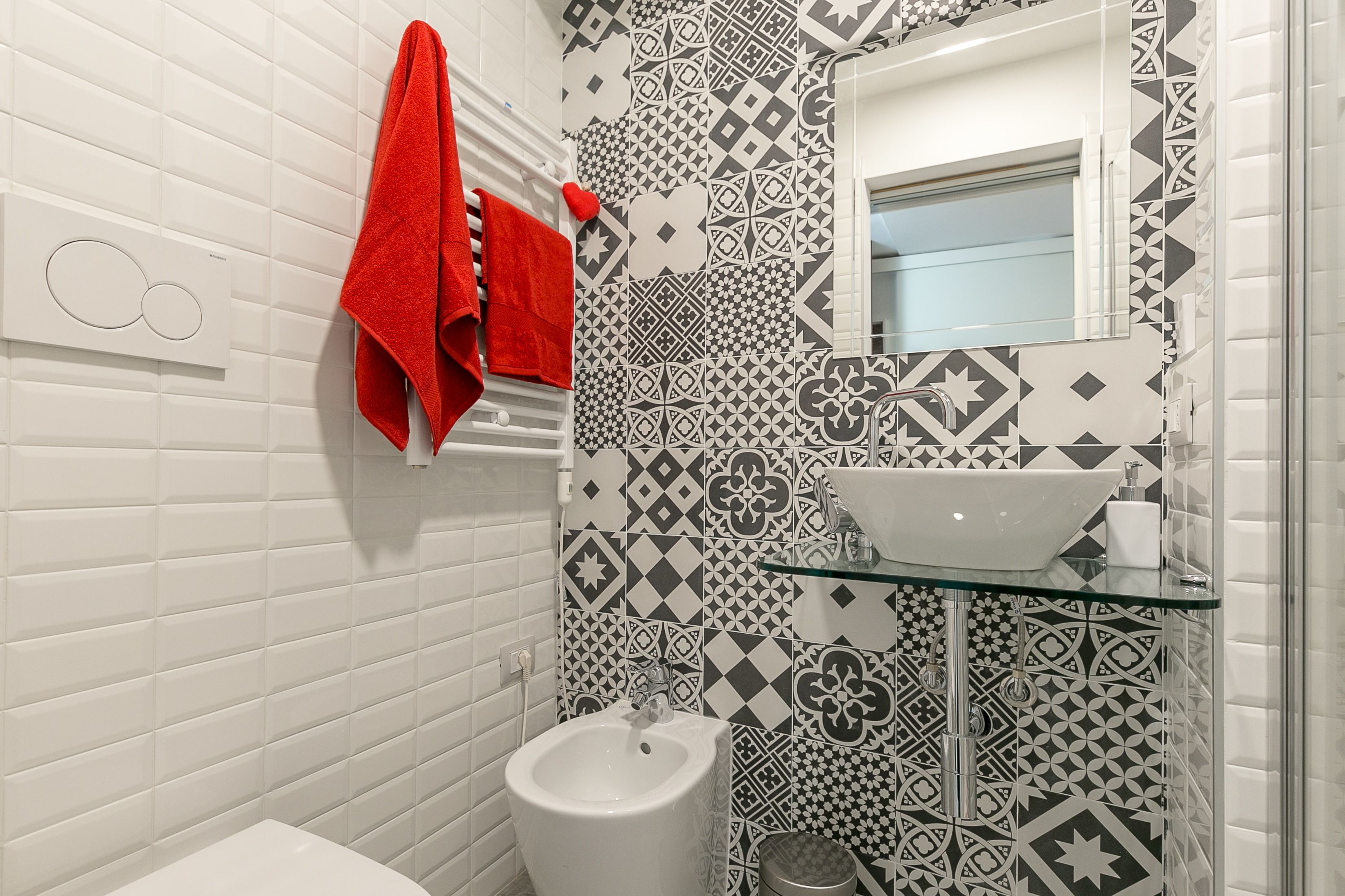 A bathroom with a bidet, a glass shelf, a white sink, and a red towel.