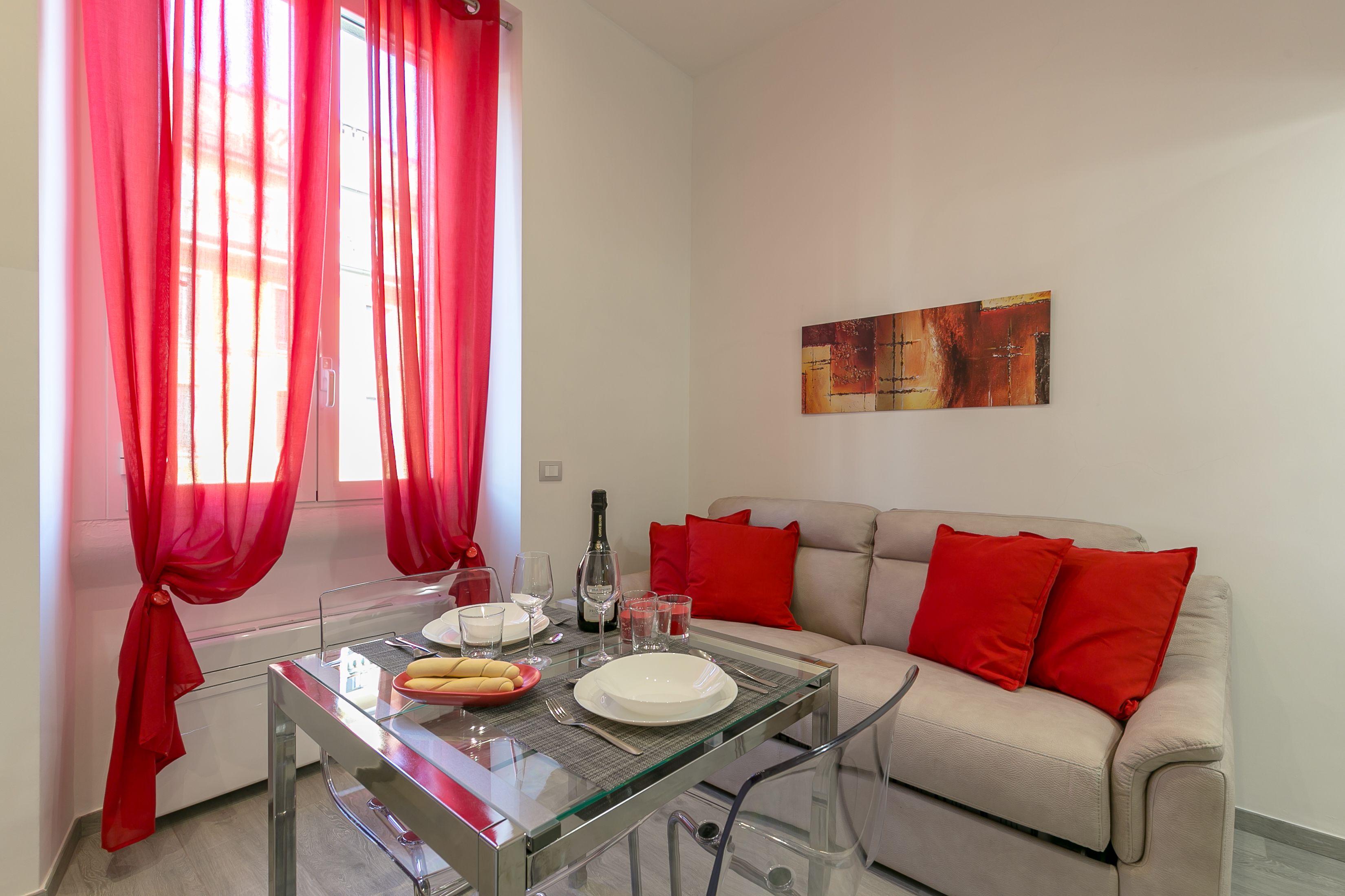 Un comedor con mesa de vidrio, cortinas rojas, un sofá con cojines rojos y un cuadro en la pared.