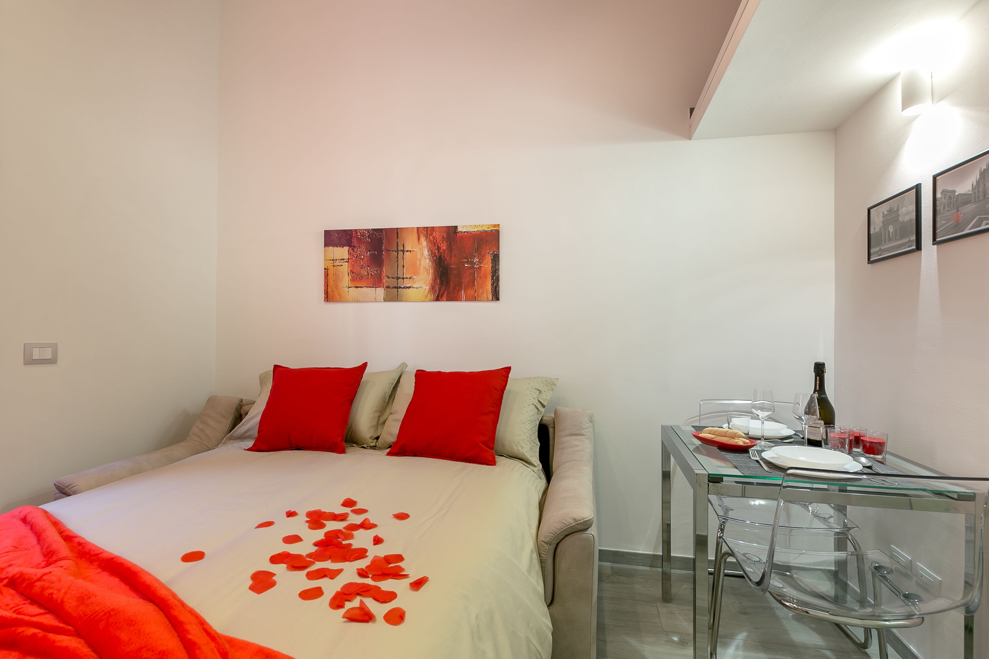 A bedroom with a bed, red pillows, a glass table with a bottle, glasses, and a plate.