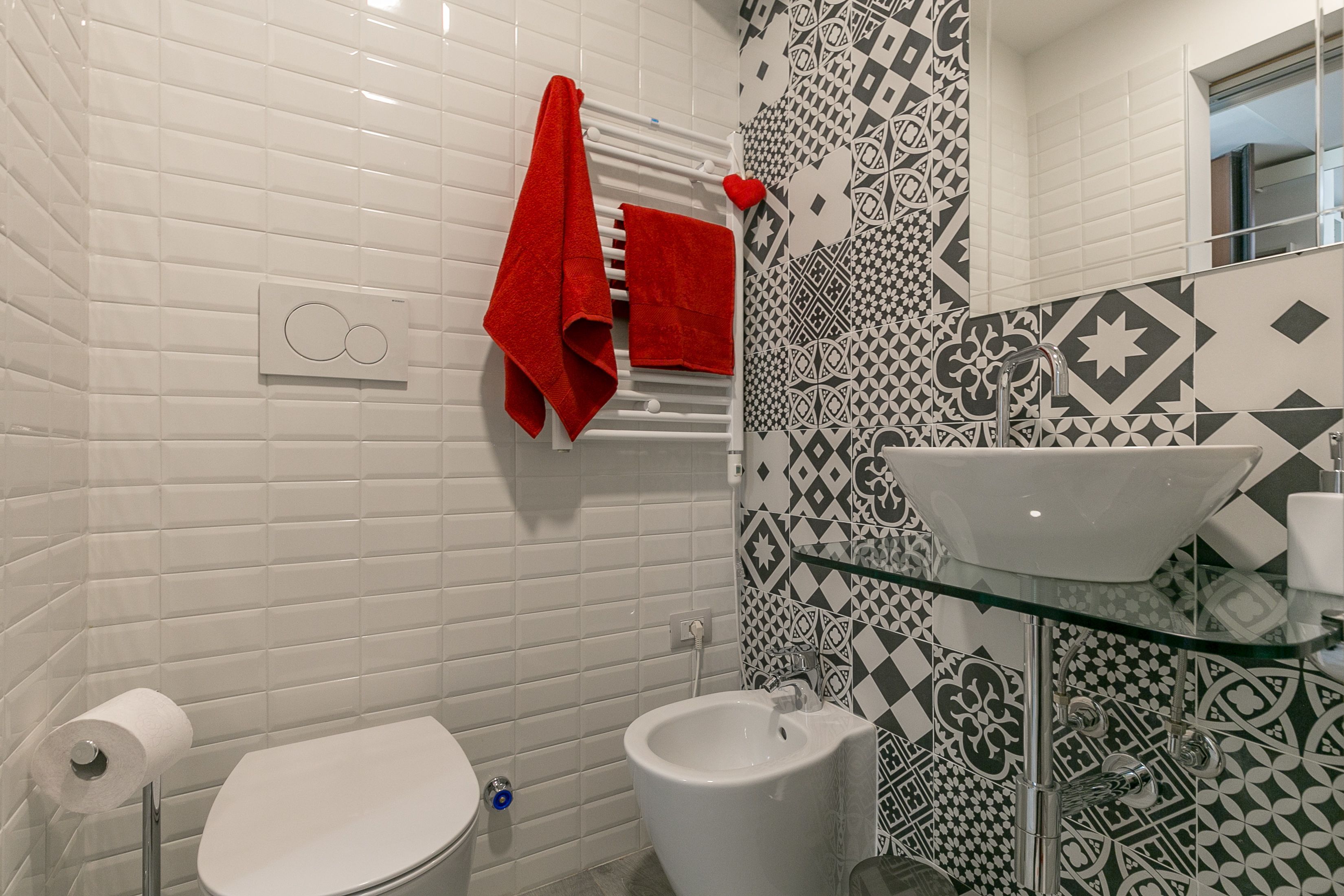A bathroom with white tiles, a toilet, a bidet, a glass sink, and a patterned backsplash.