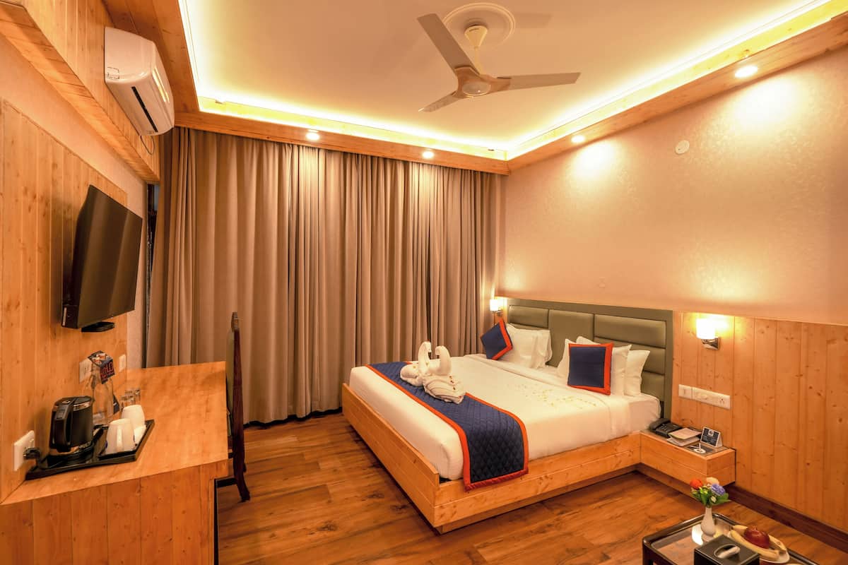 A hotel room with a wooden floor, a bed with a blue and red bedspread, a flat-screen TV mounted on the wall, a desk with a coffee maker, and a nightstand with a lamp and a vase of flowers.