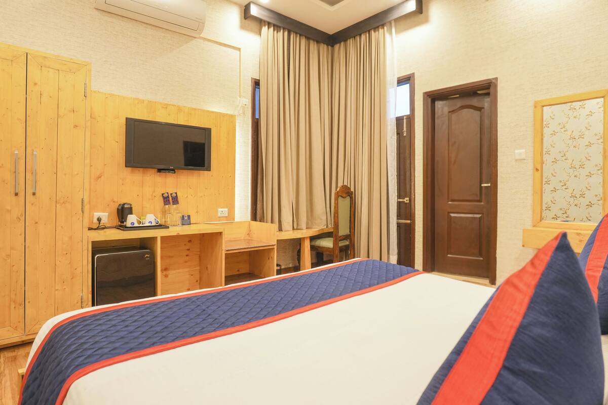 A hotel room with a bed, a TV mounted on the wall, a wooden desk with a chair, a wardrobe, and a door with a floral wallpaper.