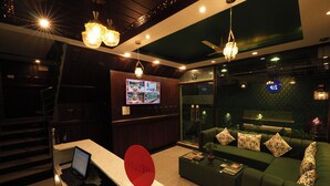 A modern lobby with a reception desk, a green sofa area, and a television screen displaying a menu. - Hotel Bella Heights (Dharamshala)