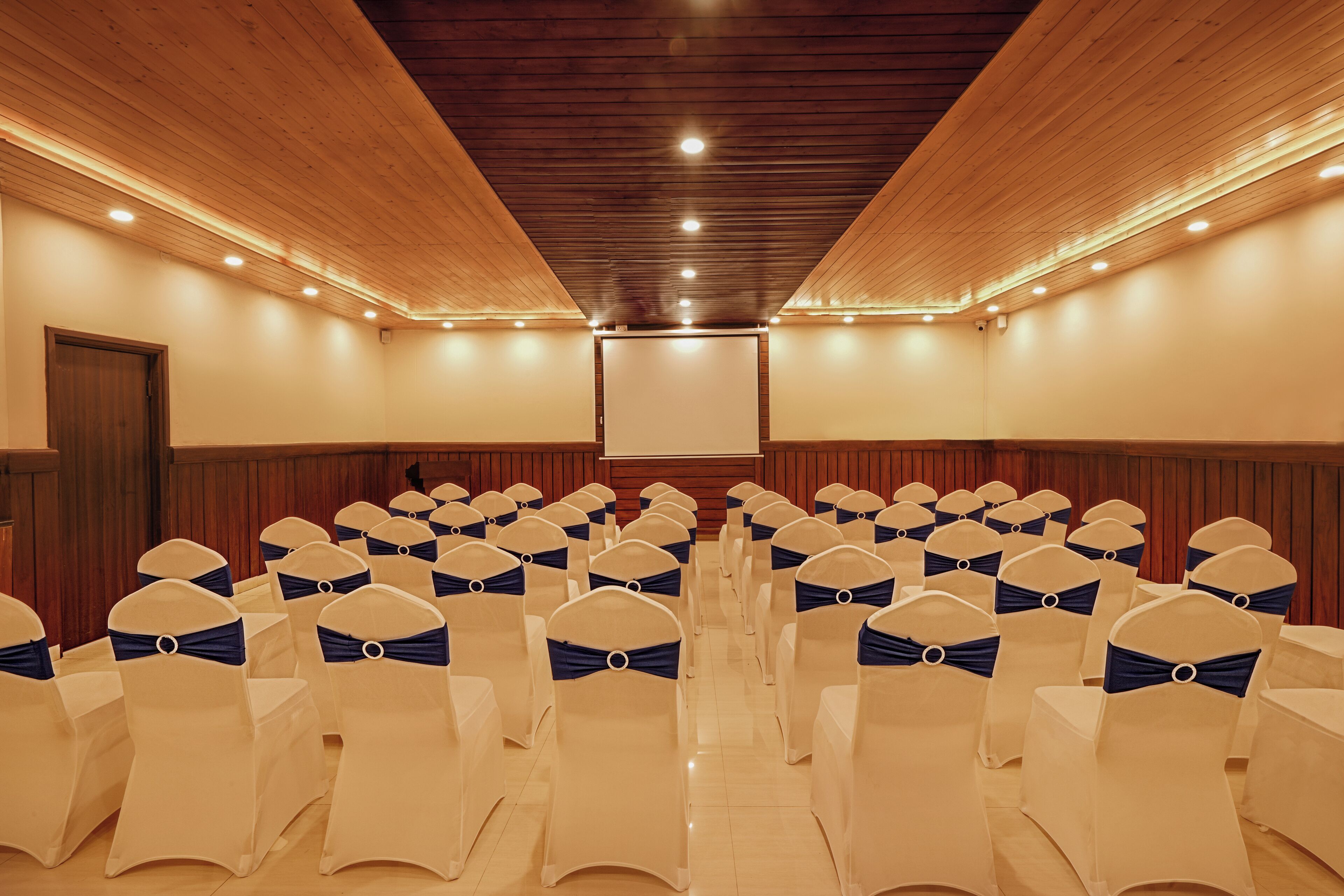 A conference room with rows of chairs, wooden panel walls, and a large screen at the front.