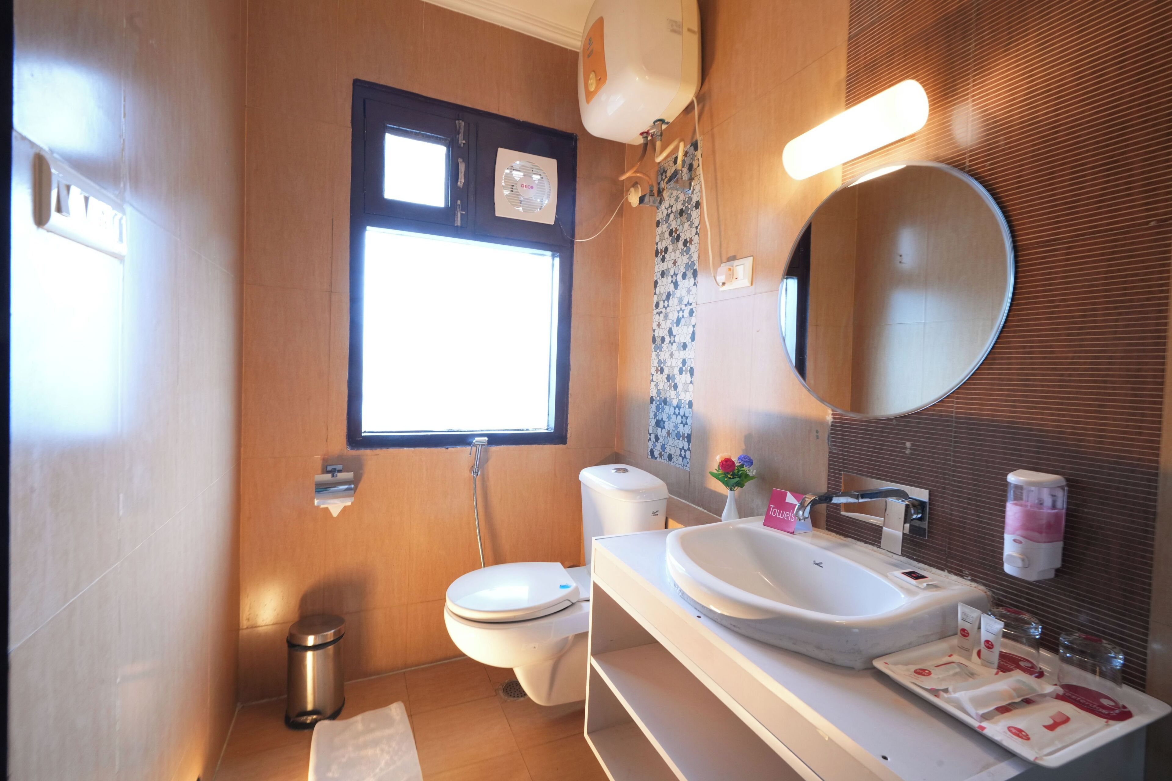 A modern bathroom with a toilet, sink, and mirror. There is a window providing natural light and a trash can.