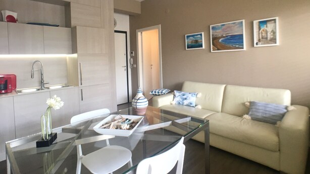 Ein modernes Wohnzimmer mit einem Glastisch, einem beigefarbenen Sofa und einer Küchenzeile mit weißer Arbeitsplatte und Schränken.
