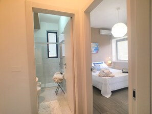 A hotel room with a shower, a bed with a white bedspread, a bedside table with a lamp, and a window with blinds. - Appartamento Mon Amour (Gallipoli)