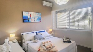 Ein Hotelzimmer mit einem großen Bett, einem Gemälde an der Wand, einem Fenster mit Jalousien und einer Deckenleuchte.