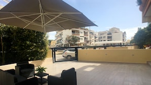 Eine Außenpatio-Terrasse mit einem großen Sonnenschirm, Korbmöbeln und Blick auf einen eingezäunten Parkplatz mit einem weißen Auto.