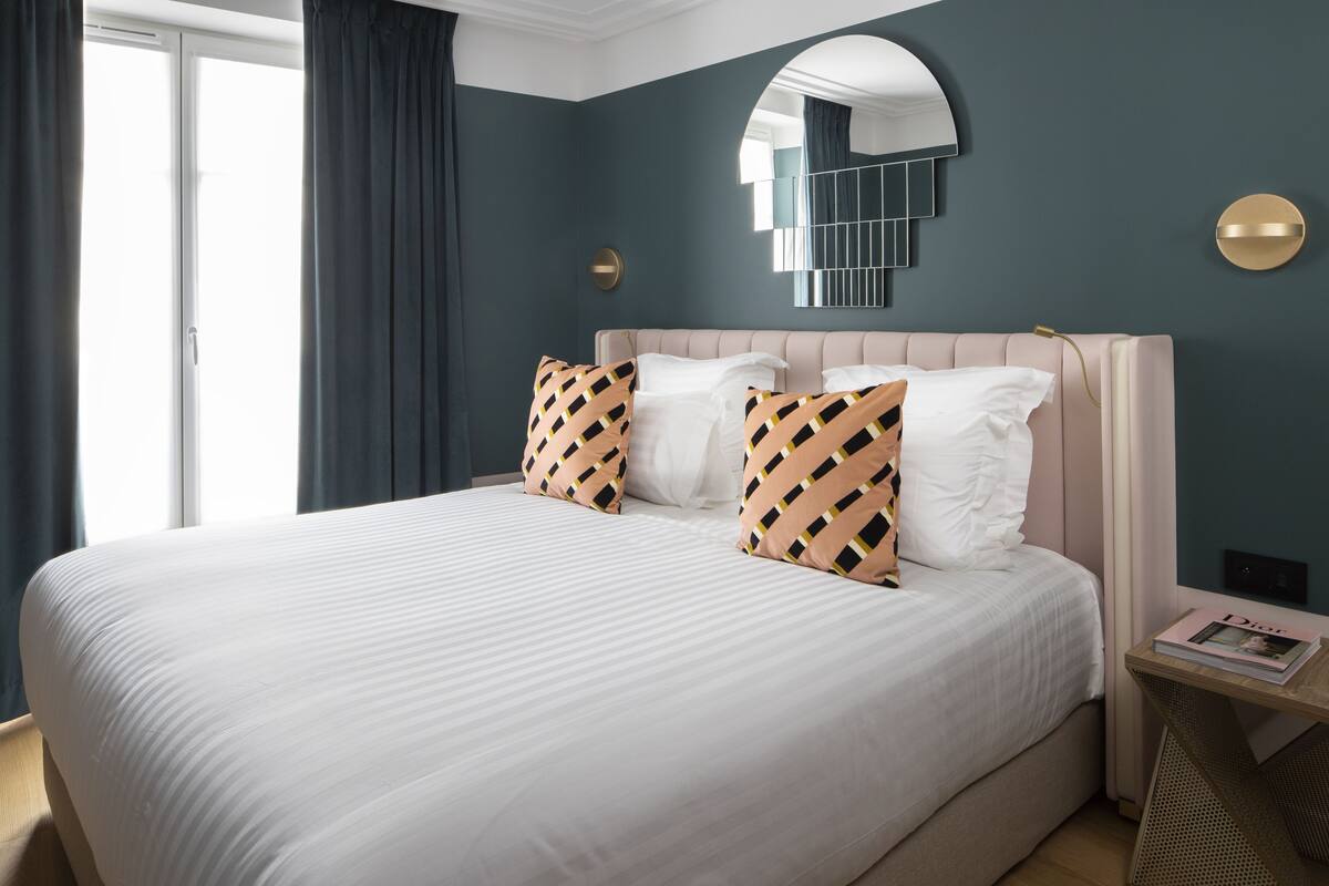 A neatly made bed with white linens and patterned pillows, a bedside table with a magazine, and a wall-mounted mirror.