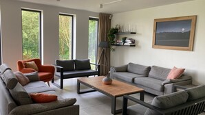 Ein modernes Wohnzimmer mit einem grauen Sofa, einem orangefarbenen Sessel und einem Holztisch. Große Fenster mit Vorhängen, eine Stehlampe und ein Bücherregal mit Büchern und Dekorationselementen.