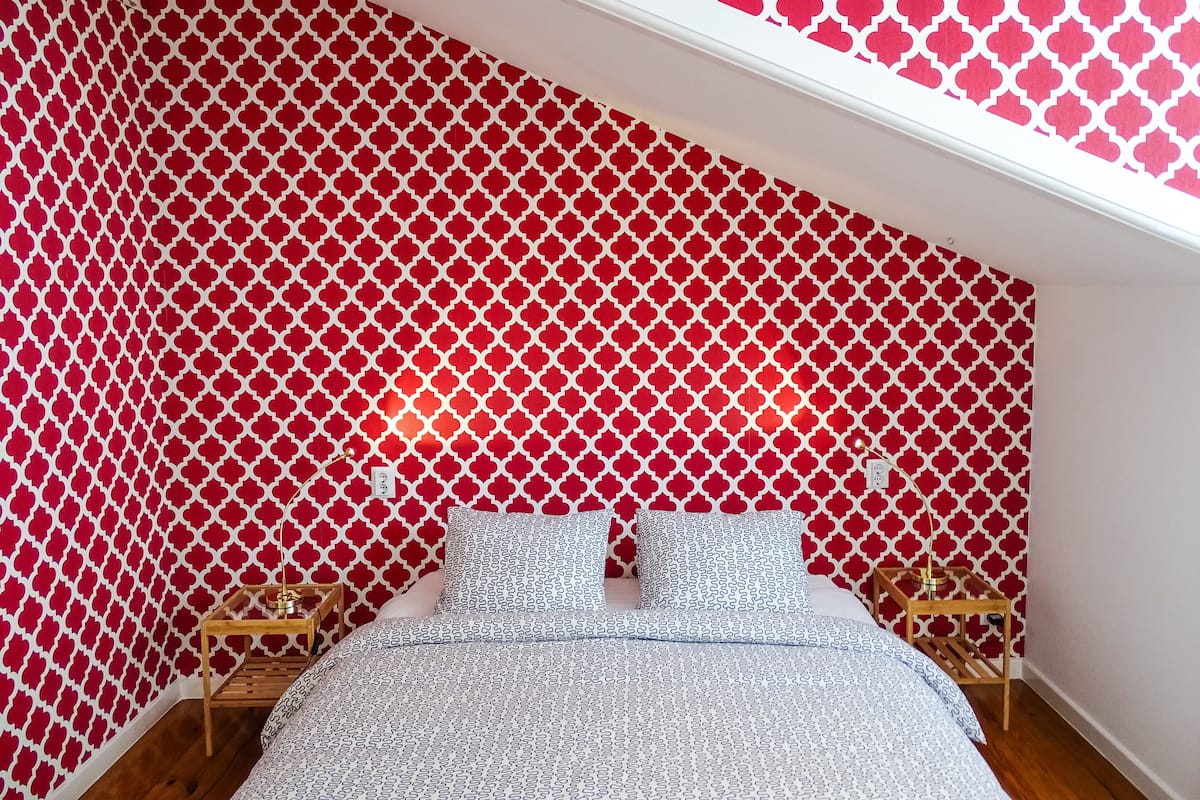 A bedroom with a patterned red and white wall, a bed with patterned bedding, and two bedside tables with lamps.