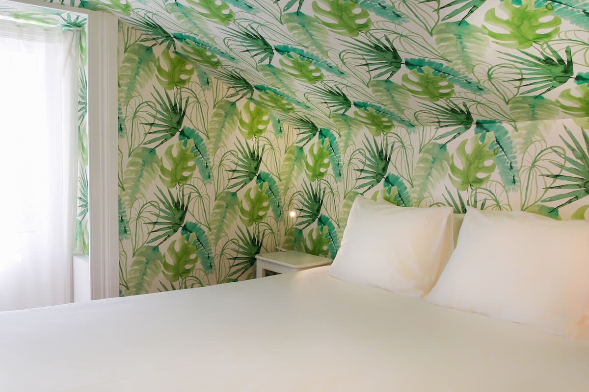 A bedroom with a bed, white pillows, and a nightstand. The walls have a green leafy wallpaper.