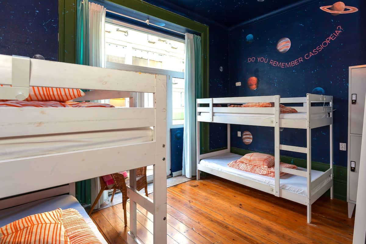 A room with bunk beds, a window with curtains, a wall with a space-themed mural, and a wooden floor.