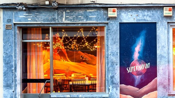 A building with a large window displaying a restaurant interior with tables and chairs, and a mural on the adjacent wall depicting a space scene with the word "SUPERNOVA". - Supernova - Hostel (Porto)