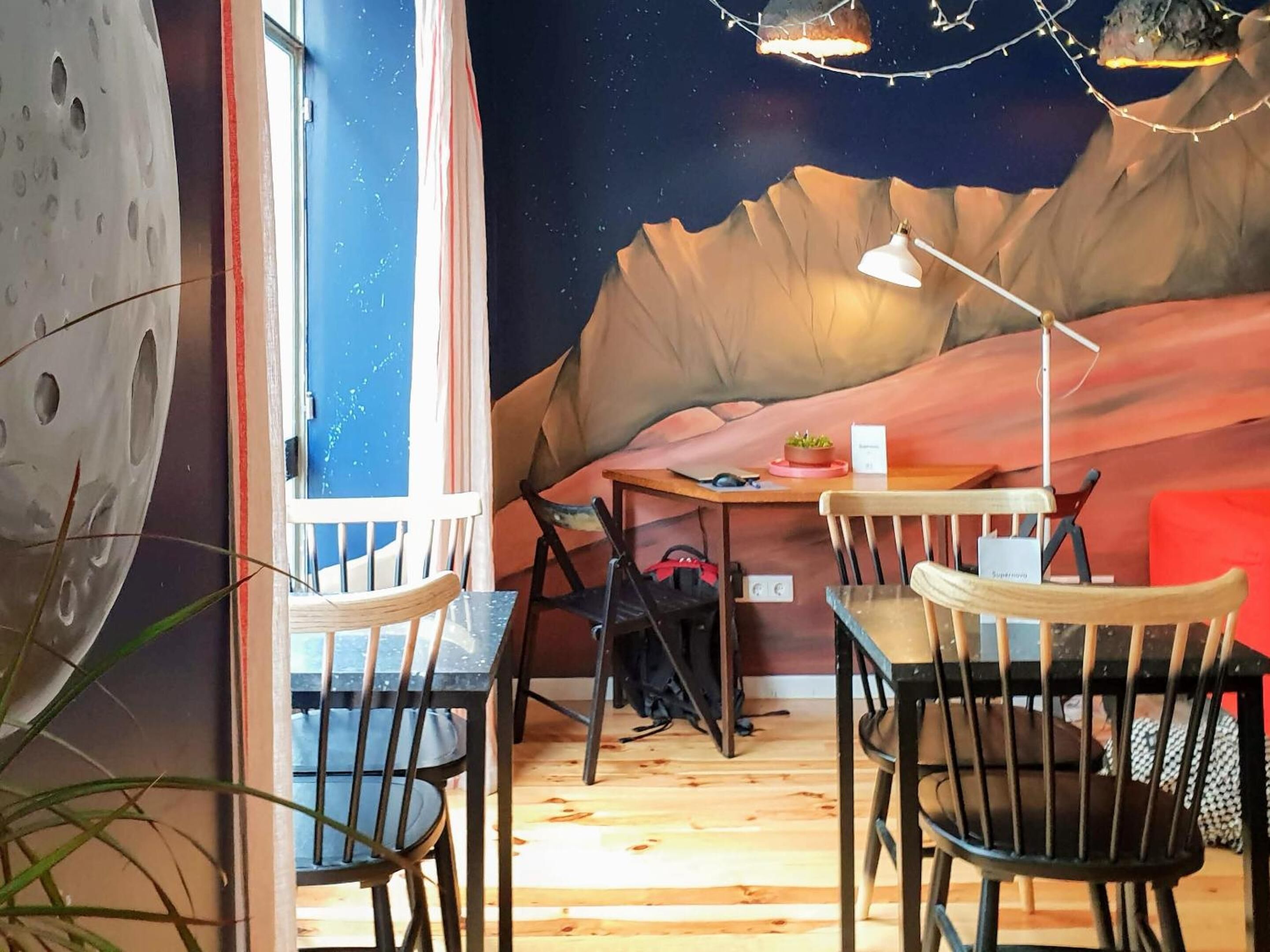 Una habitación con piso de madera, una mesa con sillas, una planta pequeña en maceta y un mural en la pared que representa un paisaje desértico.