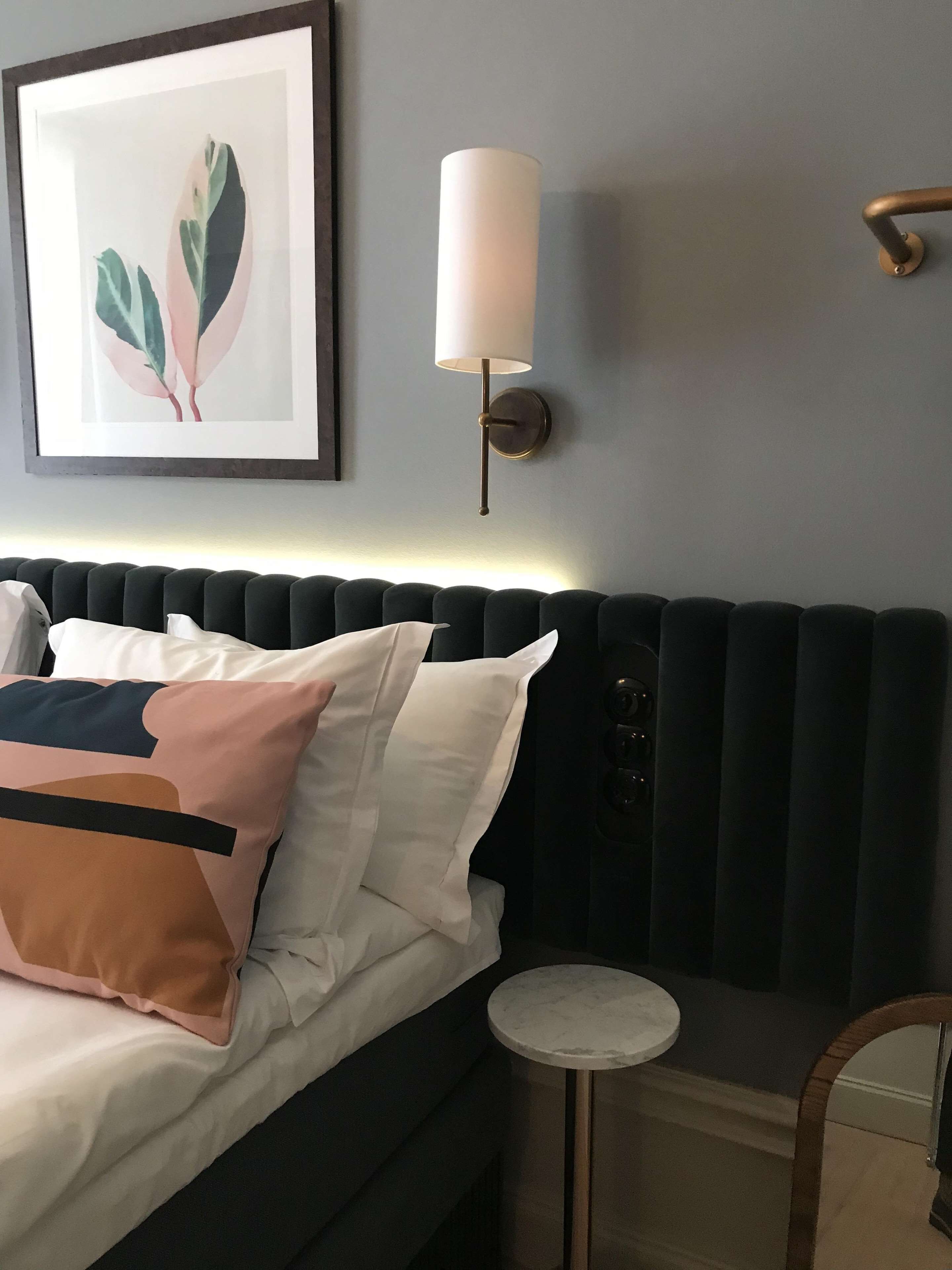 Una cama bien hecha con almohadas, una mesita redonda, una lámpara de pared y una obra de arte enmarcada en la pared.