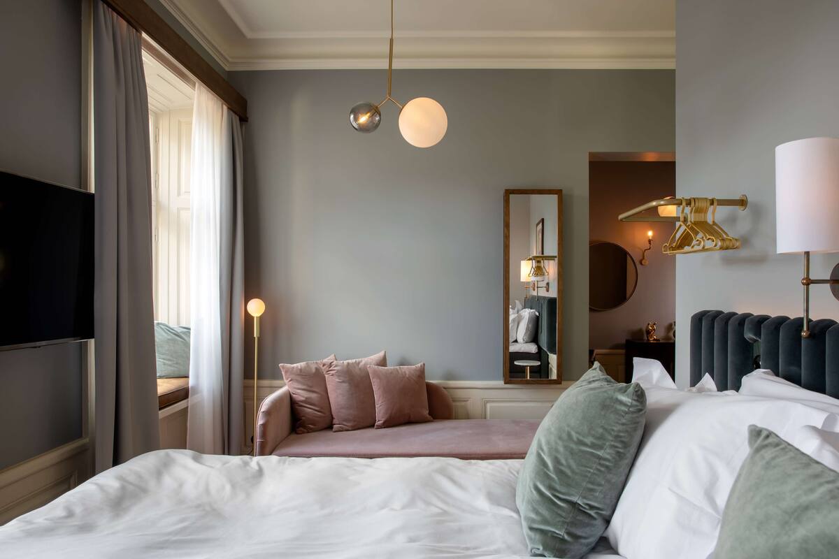 Habitación de hotel con cama, televisión, lámpara de noche, espejo y ventana con cortinas.