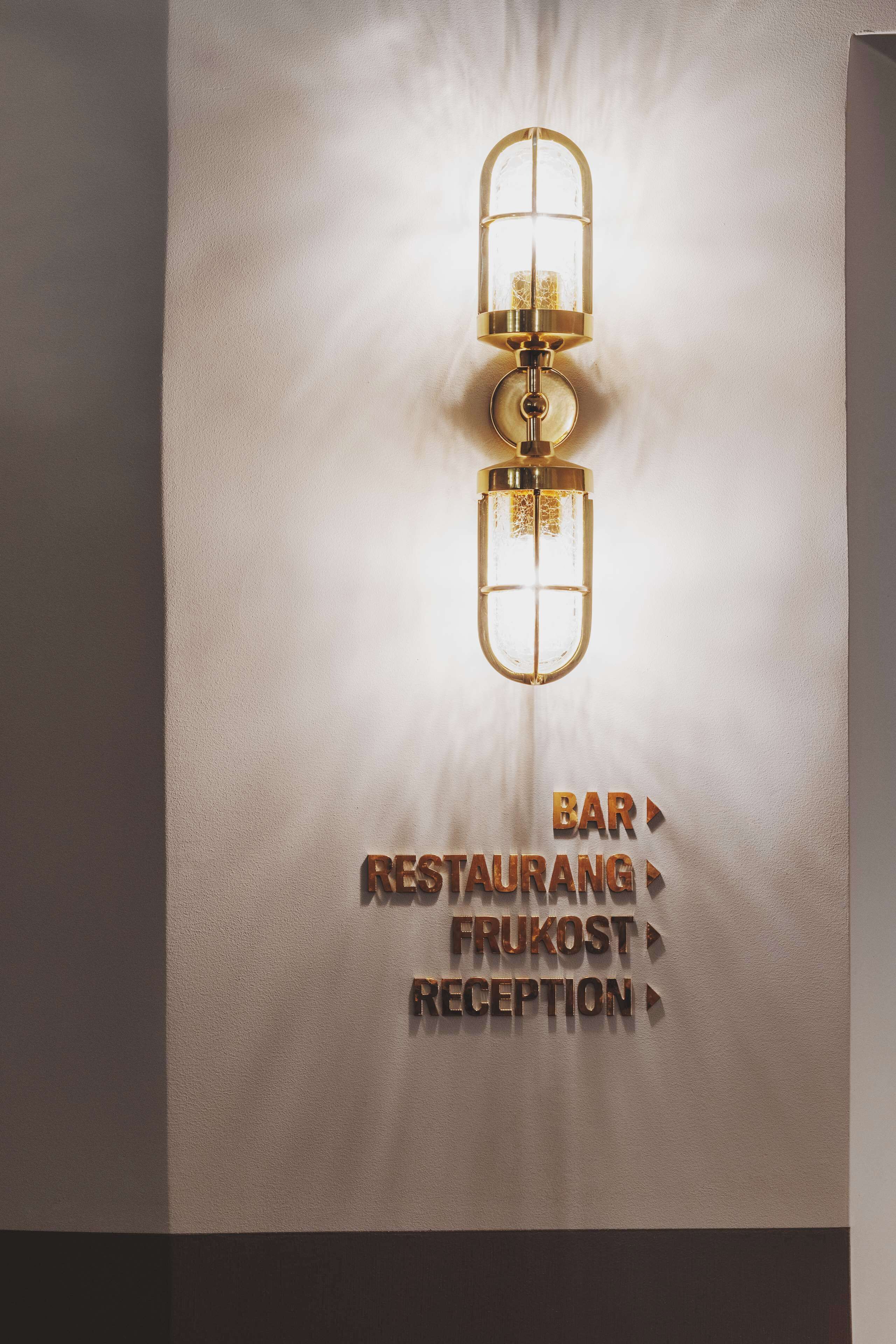 Une enseigne fixée au mur, avec un luminaire placé au-dessus, indiquant la présence d’un bar, d’un restaurant et d’un espace d’accueil.