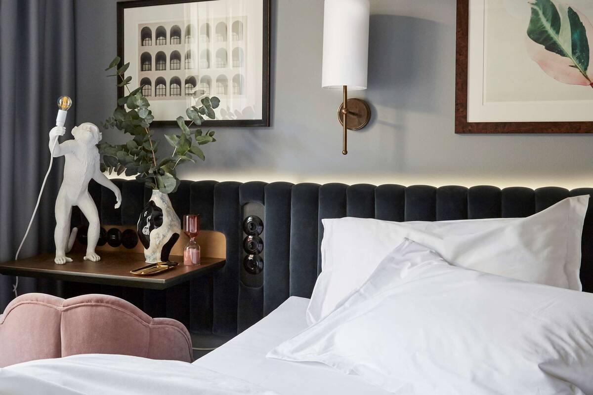 Una cama bien hecha con sábanas blancas, un cabecero tapizado oscuro, una mesita de noche con una planta y una pequeña escultura, y un cuadro en la pared.