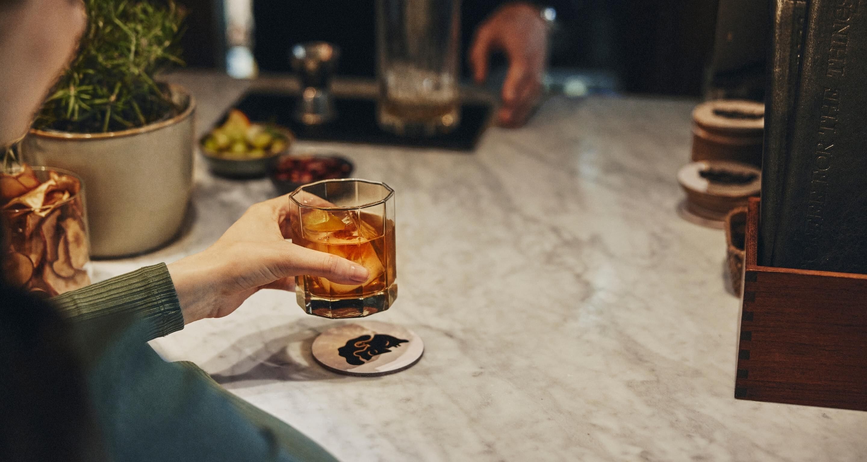 A person holding a glass of whiskey at a bar with a marble countertop, a potted plant, and a wooden cabinet.