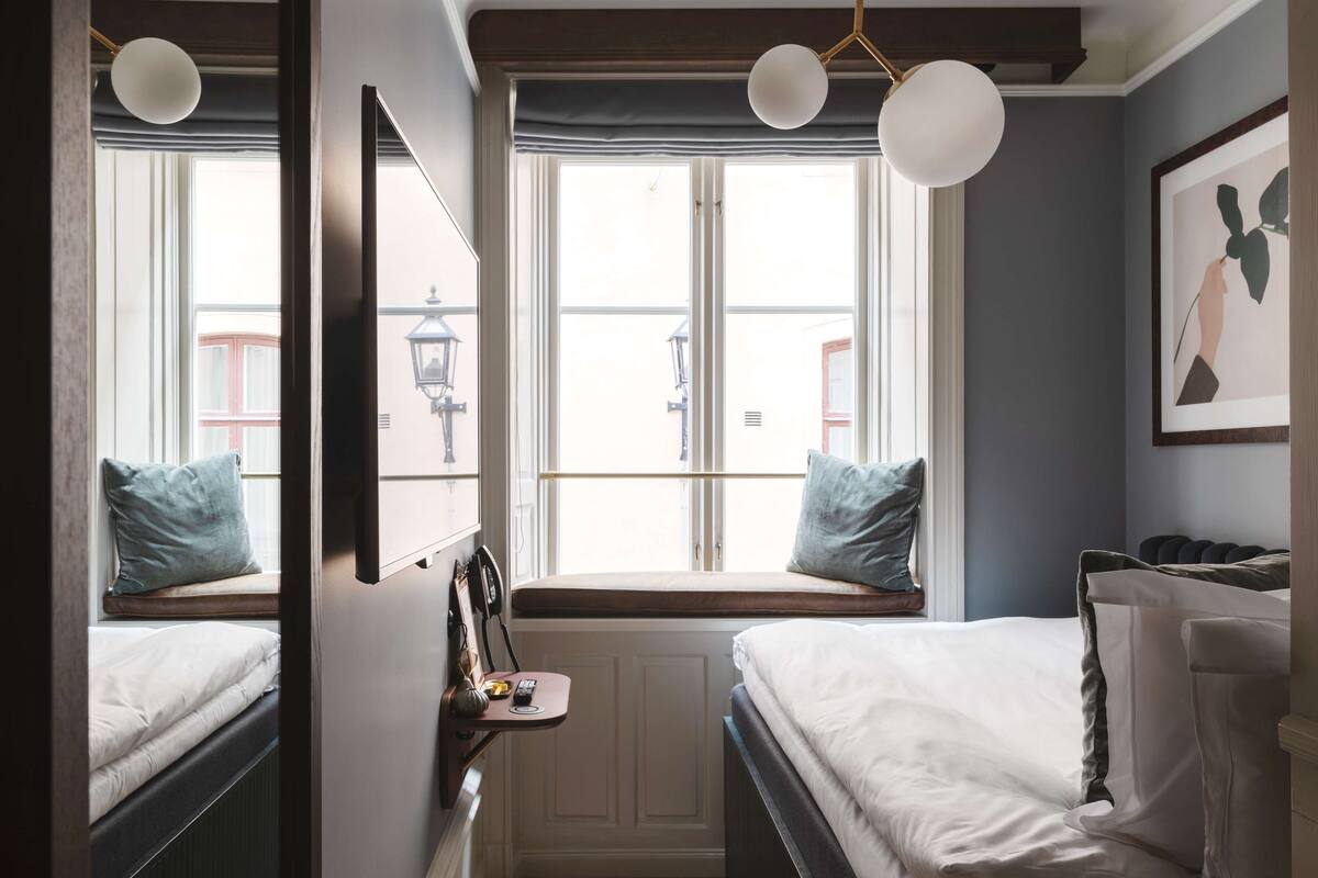 Un dormitorio con dos camas, un asiento junto a la ventana con vistas, un radiador y una lámpara de pared.