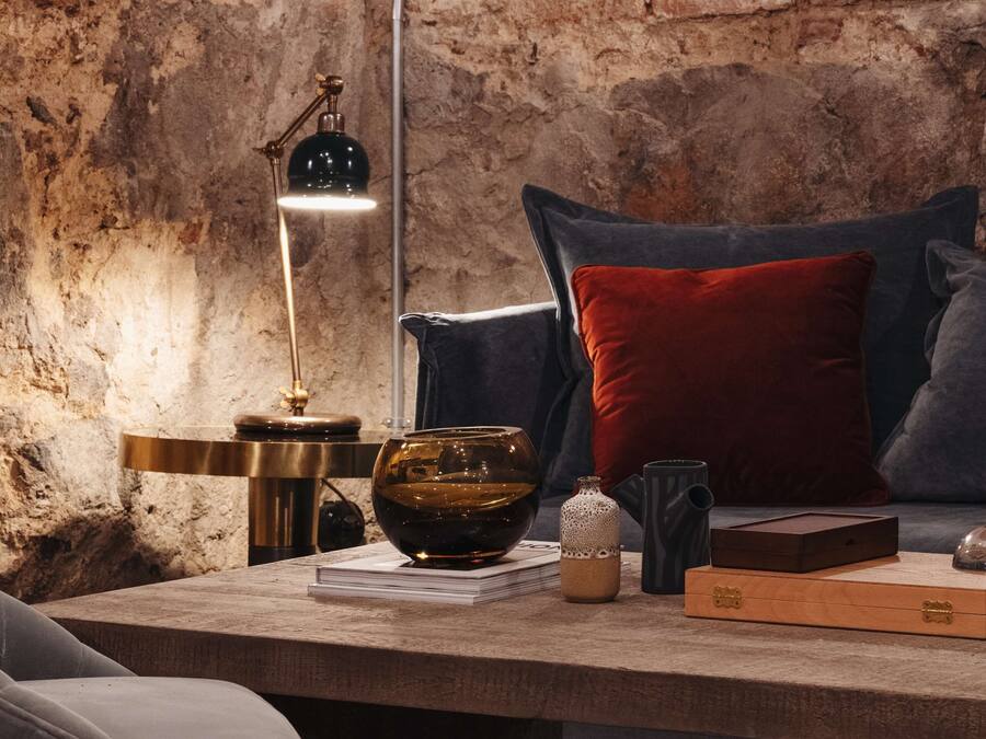 Una habitación acogedora con pared de ladrillo, una mesa de madera, una lámpara, un cuenco y un almohadón rojo.
