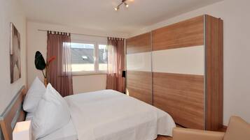 A bedroom with a bed, a wooden wardrobe, a window with curtains, and a chair.