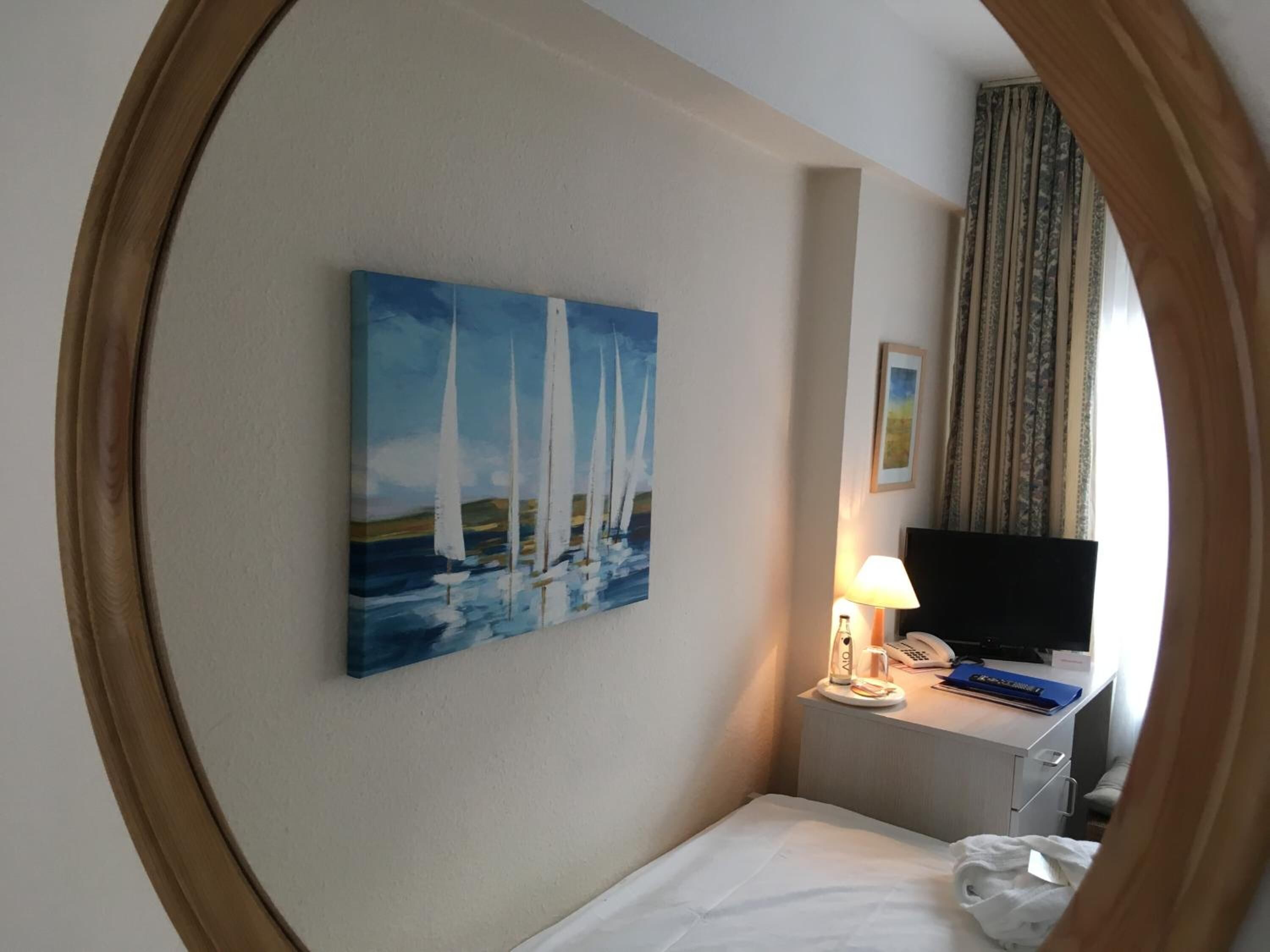 Ein Hotelzimmer mit einem Bett, einem Schreibtisch mit Lampe, einem Telefon und einem Gemälde mit Segelbooten.