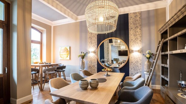 A dining area with a wooden table, grey chairs, a chandelier, and a large mirror. - Hotel Ahrbella (Bad Neuenahr-Ahrweiler)