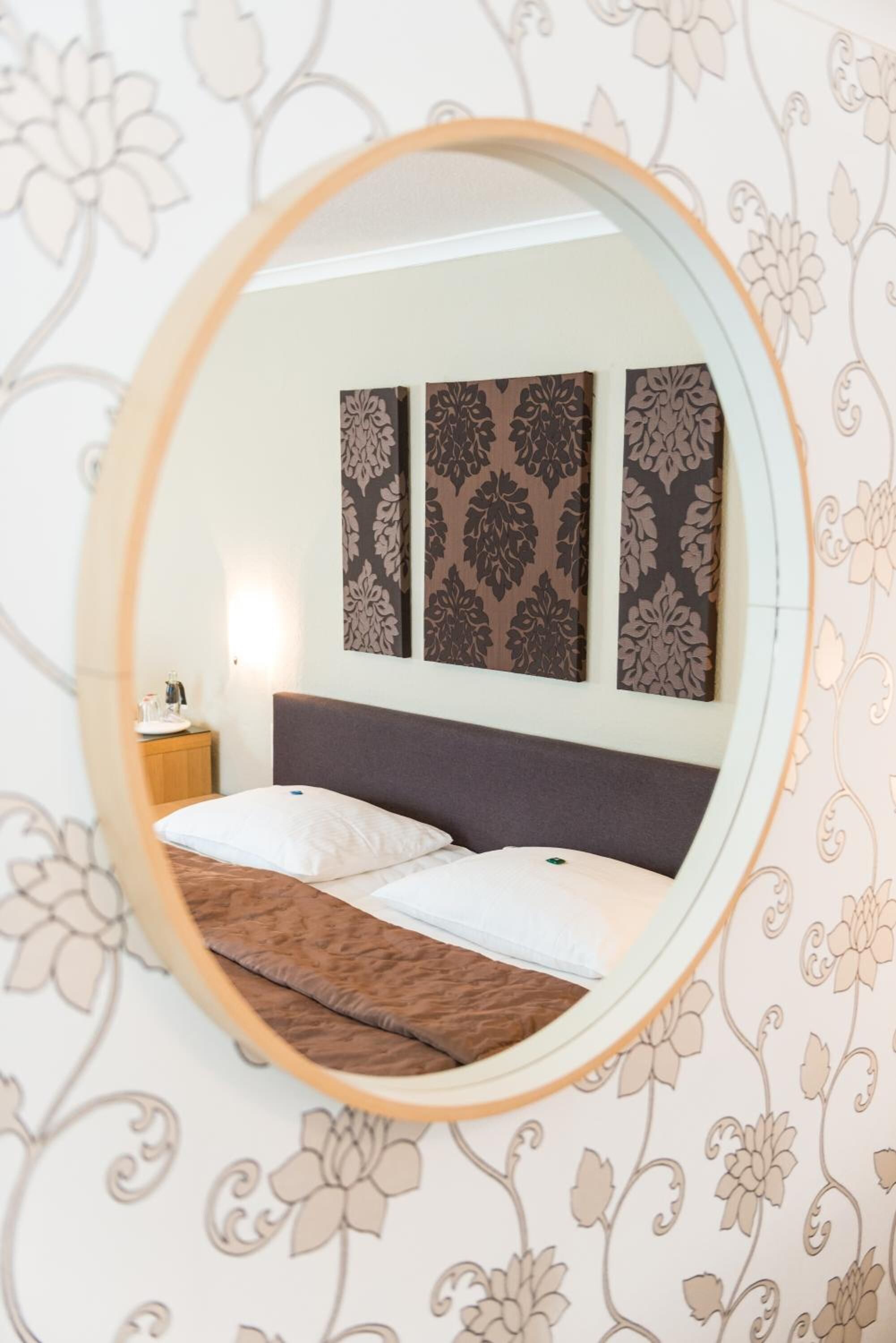 A bedroom with a round mirror reflecting a bed, a nightstand, and patterned wallpaper.