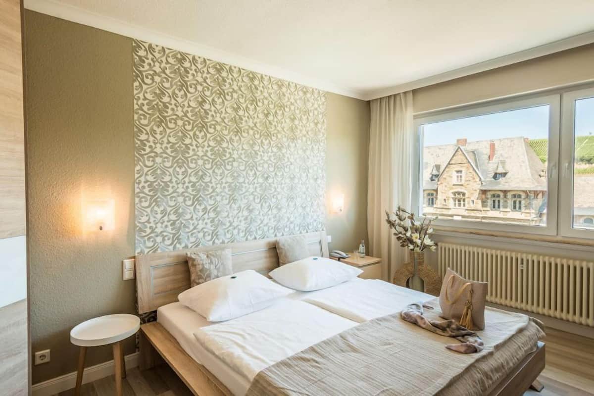 A hotel room with a bed, a nightstand, a small table, a window with a view of buildings, and a patterned wall.