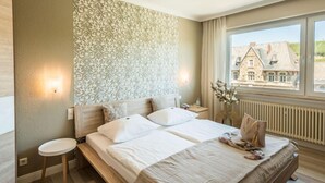 A hotel room with a bed, bedside tables, a window with a view of buildings, and a patterned wall. - Hotel Ahrbella (Bad Neuenahr-Ahrweiler)