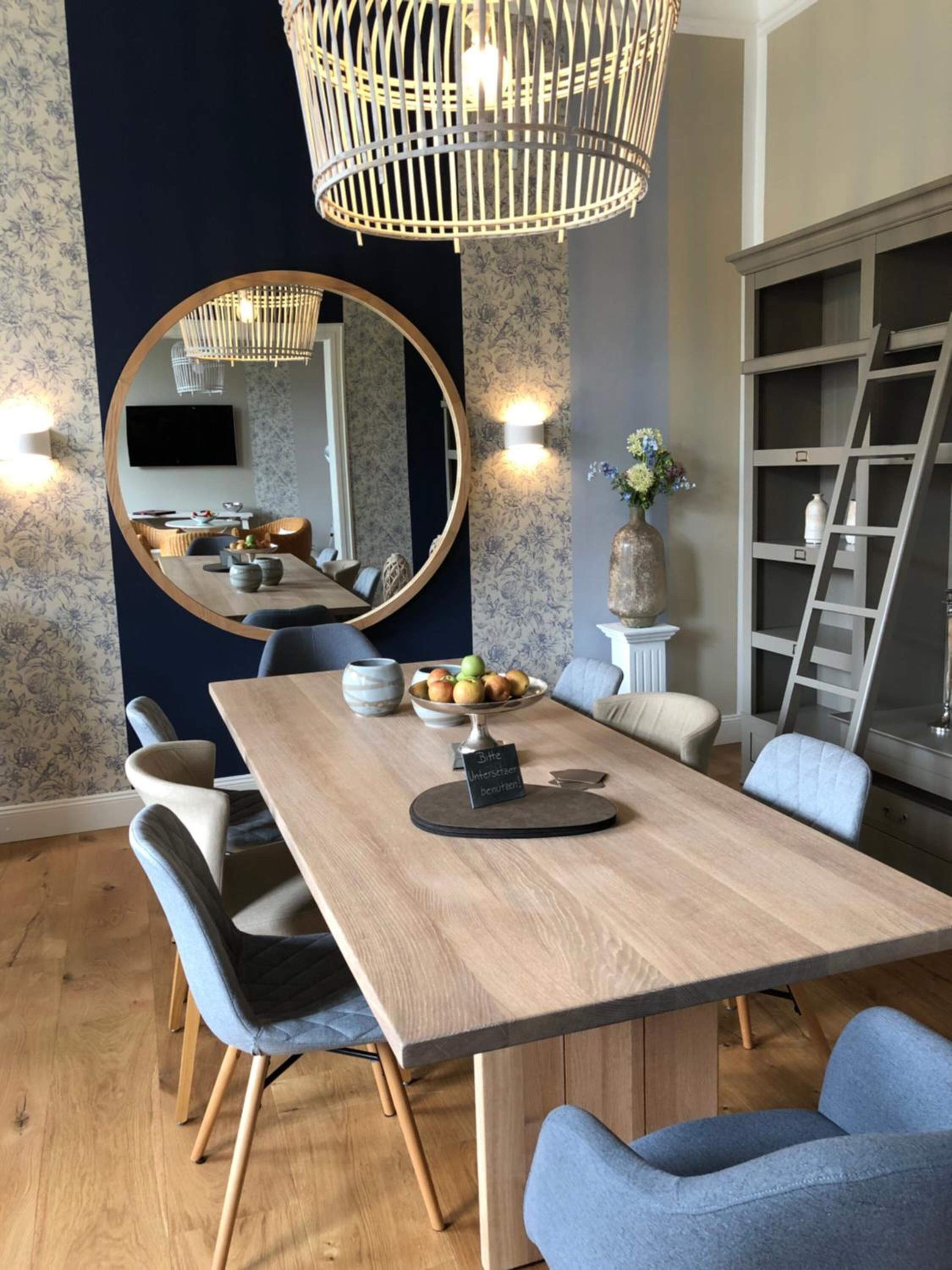 Ein Essbereich mit Holztisch, blau gepolsterten Stühlen, einem großen Spiegel und einer hängenden Leuchte.