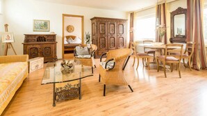 A spacious living room with a glass coffee table, wicker chairs, a wooden cabinet, and a dining table with chairs. - Hotel Ahrbella (Bad Neuenahr-Ahrweiler)