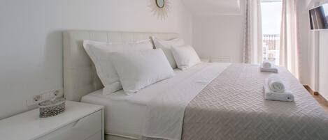 Una cama bien hecha con sábanas blancas y una colcha acolchada, una mesita de noche con un objeto decorativo y una ventana grande con cortinas.