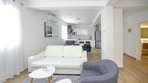 A modern living room with a white sofa, a grey armchair, and a small white table. A kitchen is visible in the background with white cabinets and a microwave. - Villa Vito (Bibinje)