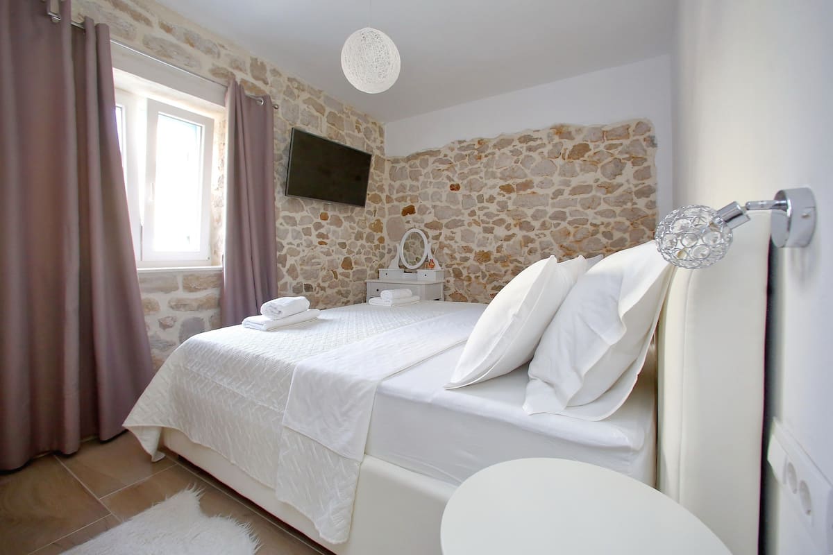 A bedroom with a stone wall, a bed with white linens, a bedside table with a mirror, and a hanging light fixture.