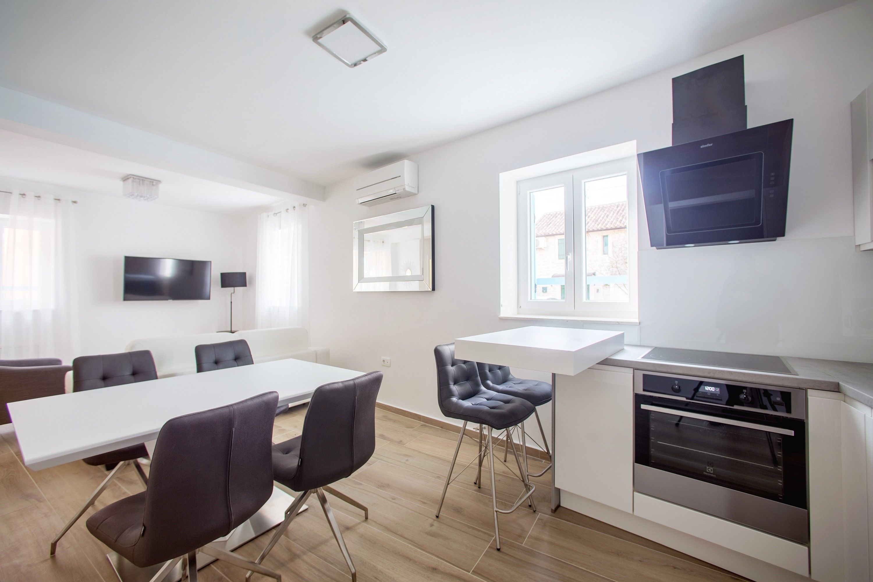 A modern kitchen with a built-in oven, a dining area with a long table and chairs, and a TV mounted on the wall.