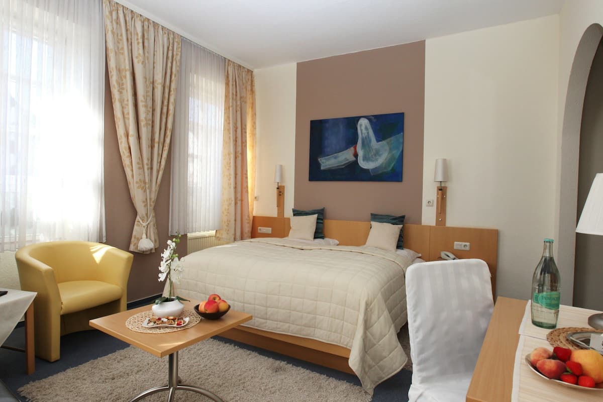 Quarto de hotel com cama, televisão, cadeira, mesa pequena com frutas e um quadro na parede.