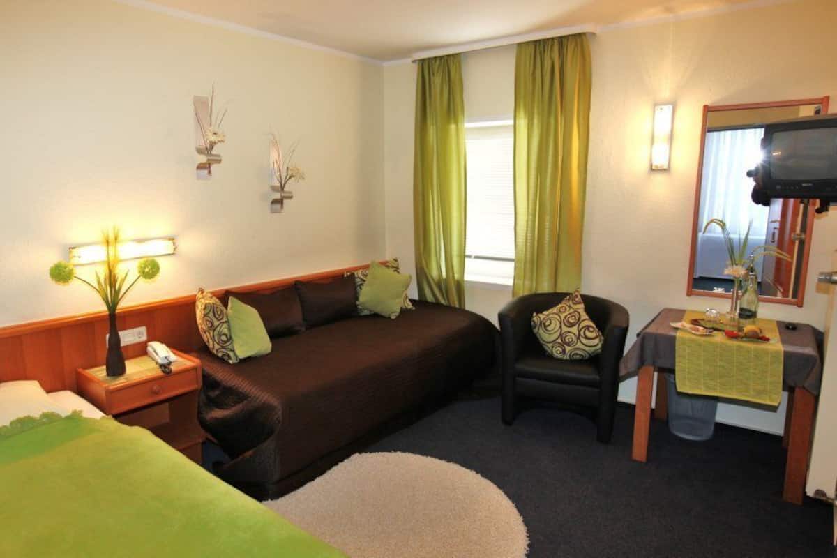 Quarto de hotel com cama, sofá, poltrona, mesa pequena, luminária, espelho e janela com cortinas.
