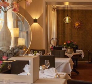 A well-lit dining area with tables set for two, a vase with flowers, and a wall-mounted light fixture. - Hotel Saarburger Hof (Saarburg)
