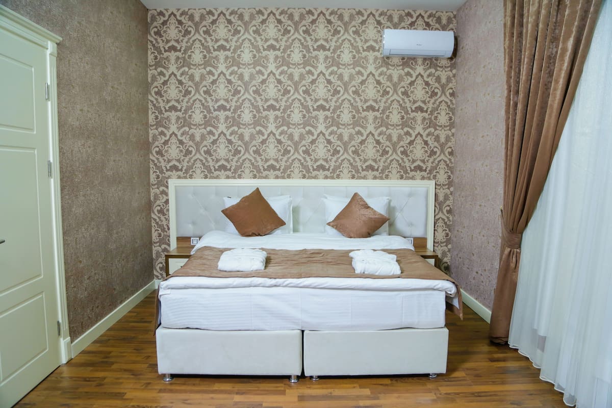 A hotel room with a bed, white curtains, a patterned wallpaper, and an air conditioning unit.